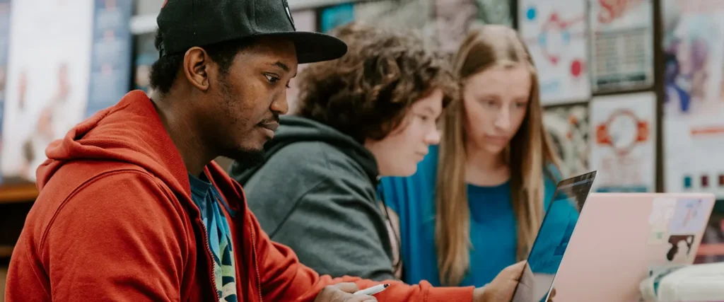 Three students sit side by side, focused on their laptops and working together in a collaborative learning environment. The student in the foreground wears a red hoodie and black cap, while the others wear blue and teal. The background features a bulletin board filled with colorful posters and announcements.