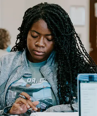 A focused student with long braided hair and a nose ring is sitting at a table, writing in a notebook. She is wearing a gray jacket and a graphic t-shirt, concentrating on her work. A blurred laptop screen is visible in the foreground, and other people are out of focus in the background, suggesting a classroom or study environment.