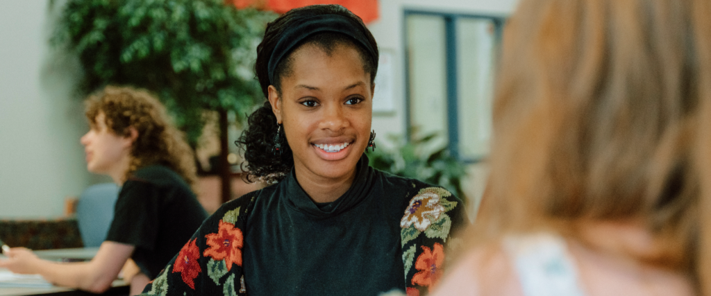 A smiling student wearing a black floral-patterned sweater and headband sits indoors, engaged in conversation with another person whose back is facing the camera. The background includes another student at a table and indoor plants, suggesting a relaxed and friendly study or common area.