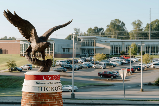 A large bronze hawk statue with wings spread sits atop a brick pedestal that reads “CVCC – Welcome to Red Hawk Nation – HICKORY.” In the background, a full parking lot and a modern academic building are visible, surrounded by green trees under a clear sky.