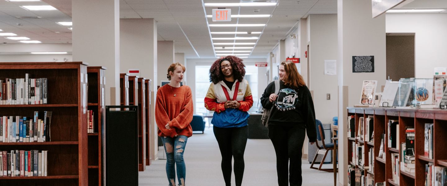 Three students walk down an aisle in a library, smiling and talking with one another. They are surrounded by bookshelves filled with books, and the well-lit hallway extends into the background. The setting conveys a welcoming and collaborative academic environment.