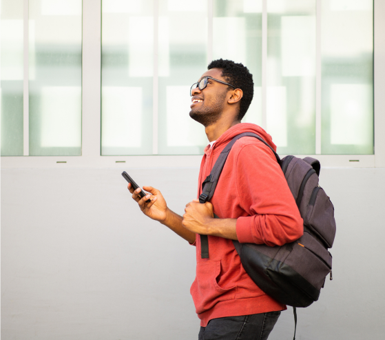 A smiling student wearing a red hoodie and backpack walks outdoors while holding a smartphone. He is looking upward with a joyful expression, and the background features large frosted glass windows set in a white wall.