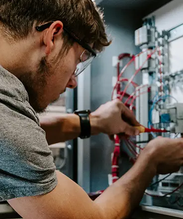 A CVCC Mechatronics student uses a screwdriver to adjust wiring in a control panel, focusing on the components of an industrial automation system.