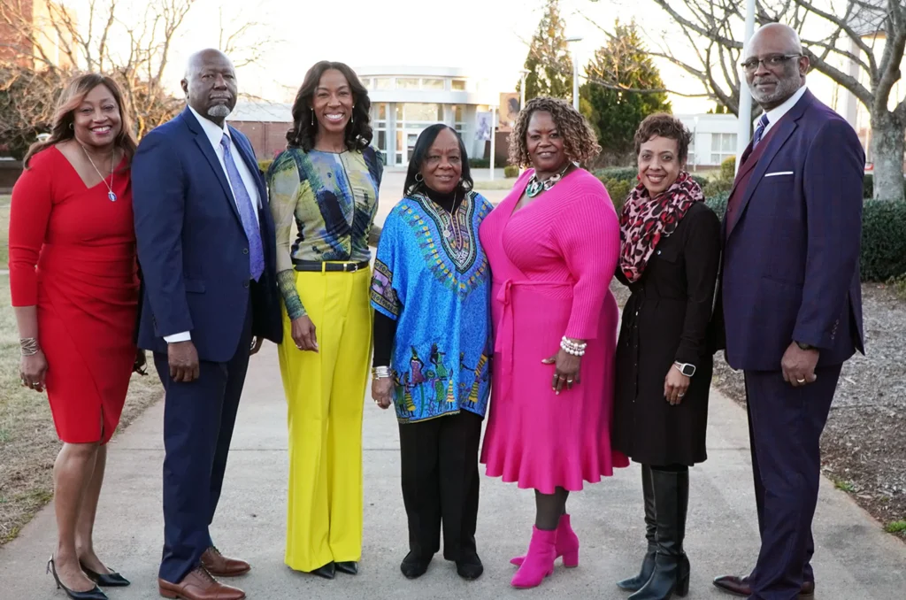 A group of seven people stands outdoors on a sidewalk, smiling at the camera. They are dressed in colorful, formal or semi-formal attire. Trees and a building can be seen in the background, suggesting a college or campus setting.