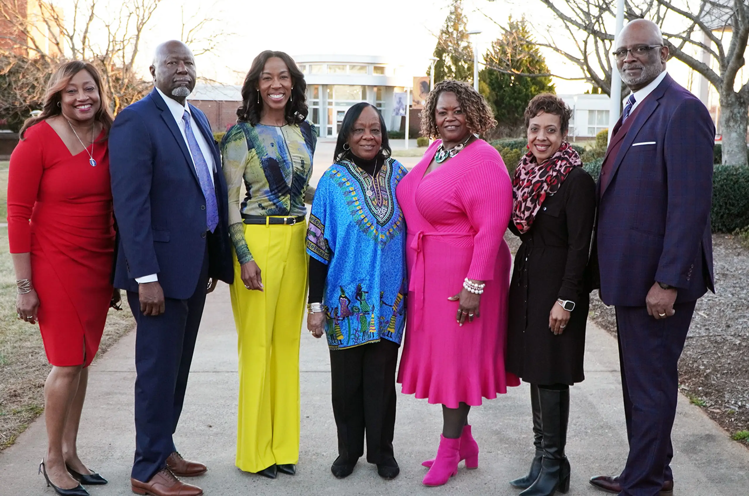 A group of seven people stands outdoors on a sidewalk, smiling at the camera. They are dressed in colorful, formal or semi-formal attire. Trees and a building can be seen in the background, suggesting a college or campus setting.