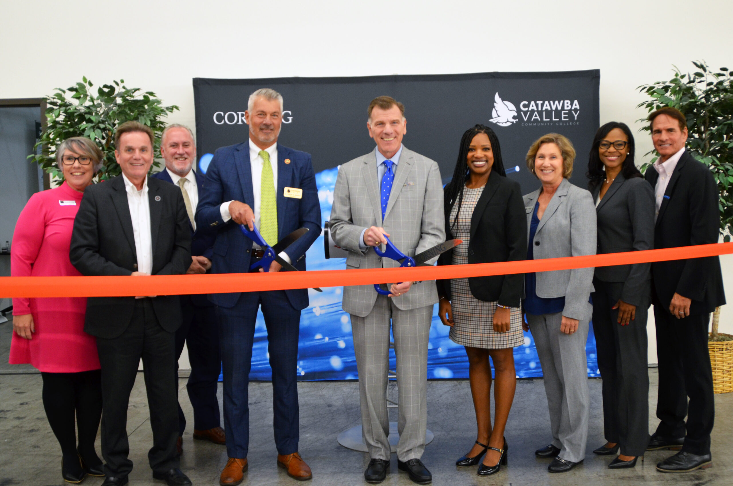 A group of ten professionally dressed individuals stand in a line, smiling, at a ribbon-cutting ceremony. Two men in the center hold oversized scissors, preparing to cut a bright red ribbon. Behind them is a backdrop displaying the Catawba Valley Community College logo and the word "Corning." Potted plants flank the scene, adding a touch of greenery.