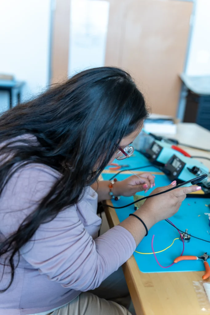A CVCC Electrical Engineering Technology student uses a soldering tool to work on wiring components at a lab bench covered with tools and colorful wires.