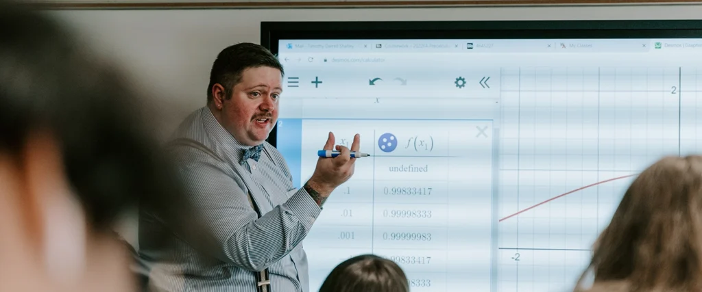 A professor, dressed in a striped shirt, bow tie, and suspenders, gestures toward a large interactive screen displaying a financial or mathematical graph. He holds a blue marker while explaining a data table and graph, likely illustrating key principles of finance, accounting, or economics. Students in the foreground attentively follow the lesson. The digital display suggests a modern, technology-integrated approach to financial education, preparing students for real-world problem-solving in accounting and finance.