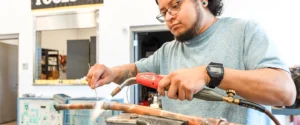 In this image, a student is engaged in a hands-on learning experience in an HVAC (Heating, Ventilation, and Air Conditioning) training lab. He is using a brazing torch and filler rod to join copper pipes, a critical skill in HVAC systems and plumbing. The workspace is equipped with tools and instructional materials, reflecting a practical, skills-based education environment. This image highlights the value of technical training programs in preparing students for in-demand careers in skilled trades.