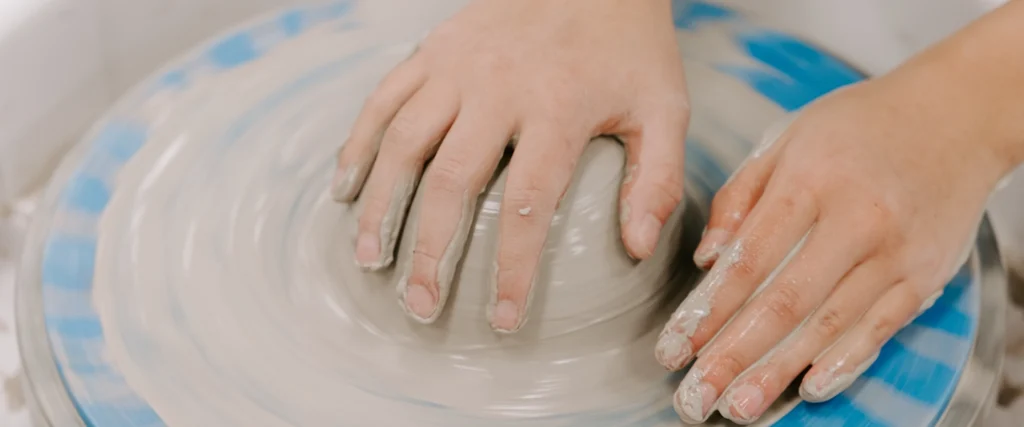 A close-up shot captures an art student’s hands covered in wet clay as they shape a spinning piece of pottery on a pottery wheel. The swirling motion of the wheel creates a mesmerizing effect, emphasizing the precision and skill required in ceramics. This hands-on experience is part of an art program that fosters creativity, craftsmanship, and self-expression through various artistic mediums.
