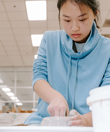 A focused art student wearing a light blue sweater carefully molds a clay bowl during a ceramics class. With hands covered in clay, she refines the shape of her piece, demonstrating patience and precision. The bright, well-lit studio environment fosters creativity, providing students with hands-on experience in pottery and sculpting techniques as part of the art program.