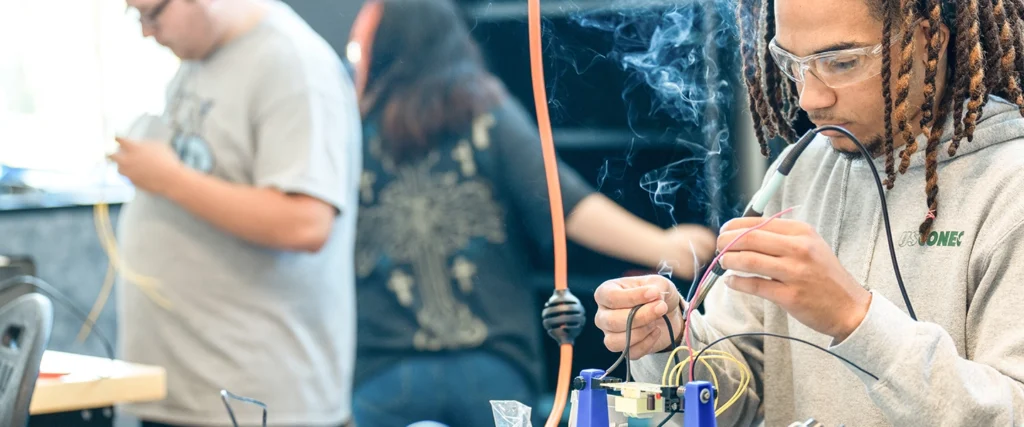 A student wearing safety glasses and a gray hoodie carefully solders wires together, with smoke rising from the connection. He is focused on assembling an electrical component, demonstrating practical skills essential for engineering studies. In the background, other students are also engaged in wiring and technical work, reinforcing a collaborative, hands-on learning environment. The lab setting, filled with tools and electrical components, showcases the applied learning approach of the Associate in Engineering program, preparing students for careers in engineering and technology fields.