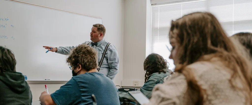A professor, dressed in a striped shirt, bow tie, and suspenders, stands at the front of a classroom, pointing to a whiteboard with handwritten notes. Students, seated at desks, are actively engaged—some taking notes with pens and others working on laptops. The classroom is well-lit, with natural light streaming through large windows, creating an inviting learning environment. The scene reflects an interactive business administration class, where students develop analytical and communication skills essential for future careers in business and management.