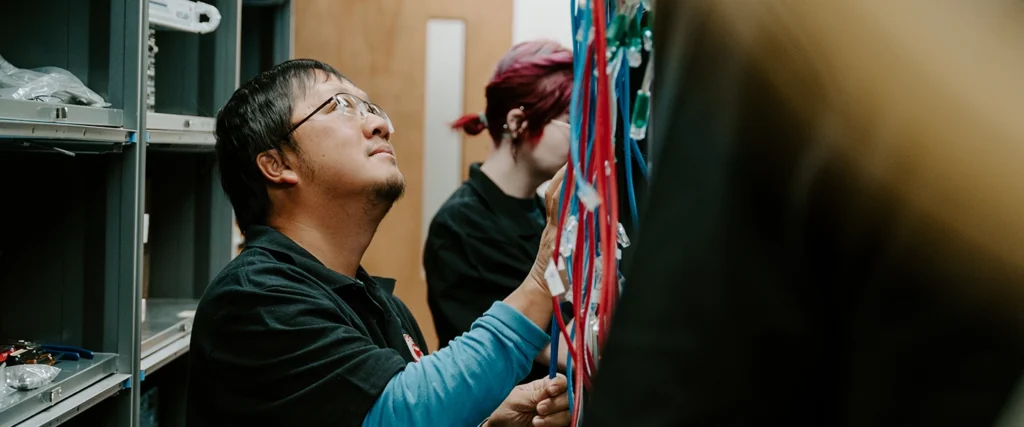 A student wearing glasses and a black polo shirt carefully works with a network wiring panel, inspecting and organizing colorful Ethernet cables. Another student with red hair is focused on a similar task in the background, demonstrating collaboration in a lab environment. Surrounding them are shelves stocked with technical tools and equipment, reinforcing the hands-on, problem-solving nature of the Computer Engineering Technology program. This learning environment prepares students for careers in networking, hardware maintenance, and IT infrastructure.