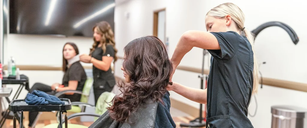 A young hairstylist with blonde hair, dressed in a black uniform, styles a client’s long, wavy brown hair in a professional training setting. The client sits in a salon chair, draped in a black cape. In the background, two other hairstylists, also in black uniforms, are attending to another client, who is having a neck strip applied. The salon is well-lit, with styling tools and equipment neatly arranged on nearby stations, indicating a hands-on learning environment.