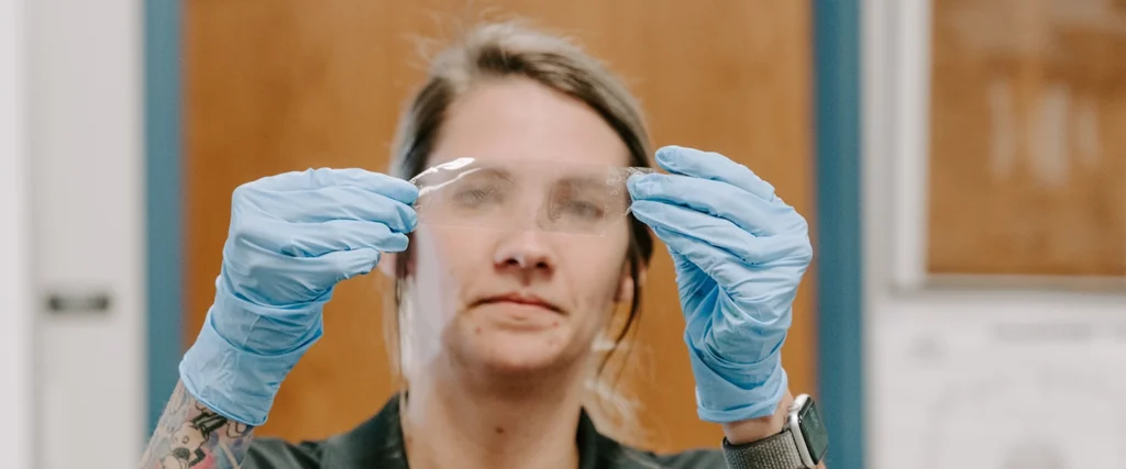 A woman wearing blue latex gloves holds up a transparent plastic sheet with a visible fingerprint, carefully inspecting it. She has a focused expression, and her attire, along with her gloves, suggests a forensic or laboratory setting. Her arms feature tattoos, and she wears a smartwatch on her wrist. The background includes a wooden door and a bulletin board, indicating a classroom or lab environment, possibly for forensic science training or analysis.