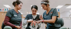 Three Dental Hygiene students, dressed in matching scrubs with name tags, work together on a dental training mannequin. They use dental instruments to practice techniques in a professional learning environment that simulates a real dental clinic. The setting includes dental chairs, equipment, and educational materials, emphasizing hands-on training. This program prepares students for careers in oral healthcare by teaching preventive care, patient education, and clinical procedures essential for maintaining dental health.