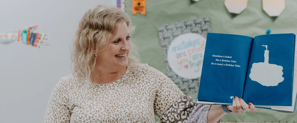 A smiling woman with wavy blonde hair, wearing a leopard-print blouse, holds up an open picture book while reading aloud. The book features a blue page with white text and an abstract illustration resembling a birthday cake. The text reads, "Sometimes it looked like a Birthday Cake. But it wasn’t a Birthday Cake." She appears engaged and happy, looking towards her audience. Behind her, a decorated classroom bulletin board displays colorful artwork and an encouraging message that says, "Mistakes are proof you are trying." The environment suggests an engaging and supportive learning space for young students.