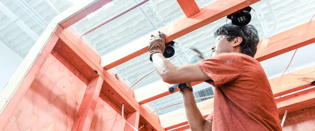 A student wearing safety glasses and work gloves carefully secures an electrical component to a wooden frame while installing wiring. He uses a hammer to fasten elements in place, demonstrating practical skills essential for electrical systems work. The workshop setting, with exposed wooden beams and structured wiring pathways, provides a realistic training experience for students preparing for careers in electrical installation, maintenance, and repair.