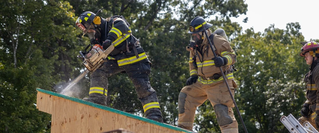 A group of firefighters in full protective gear, including helmets and oxygen tanks, work together on a slanted rooftop. One firefighter in black gear is using a chainsaw to cut into the wooden structure, with sawdust spraying into the air. Another firefighter in tan gear, wearing a breathing mask, stands nearby holding a tool, while a third firefighter in a red helmet climbs a ladder to assist. The background consists of lush green trees under a bright sky, suggesting an outdoor training exercise or an emergency response in progress.