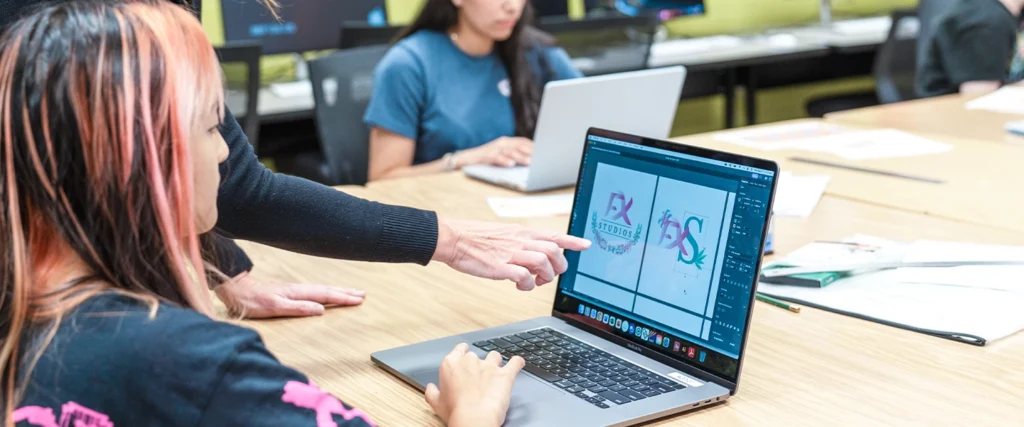 A student with pink and black hair works on a logo design in a graphic design software program on a laptop. An instructor, dressed in black, points at the screen, providing guidance and critique. The screen displays two versions of a logo for "FX Studios" with colorful, artistic elements. Other students in the background are also engaged in design work, using laptops and digital tools. The classroom setting emphasizes creativity, technical skill development, and collaboration, key aspects of a Graphic Design program.