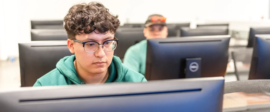 A student wearing glasses and a green hoodie concentrates on his computer screen in a modern cybersecurity lab. Rows of Dell desktop monitors fill the room, creating a structured learning environment. Another student in the background, also in a green hoodie, is engaged in his work, emphasizing the collaborative yet focused nature of the program. The setting highlights the technical skills and problem-solving mindset required for careers in cybersecurity, including ethical hacking, network defense, and data security.