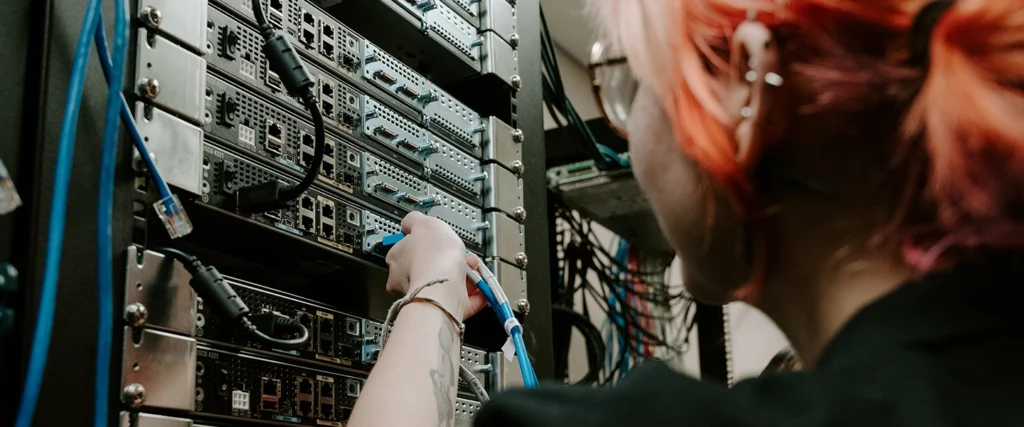 A student with vibrant pink and orange hair, wearing safety glasses, carefully plugs a blue Ethernet cable into a server rack filled with networking hardware. The dimly lit environment, filled with neatly arranged cables and multiple server units, resembles a real-world data center or IT lab. The student's focused posture highlights the technical skills necessary for careers in IT support, network administration, and cybersecurity. This hands-on learning experience prepares students to manage and maintain complex information systems.