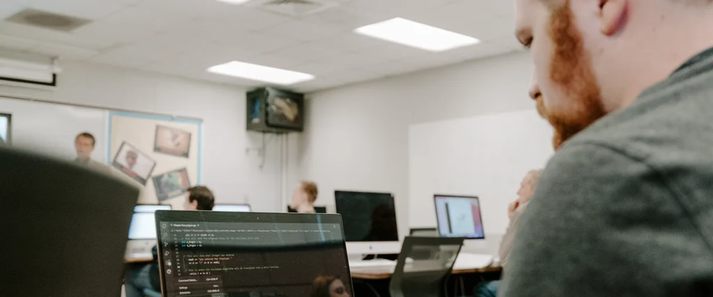 A student focuses on his laptop screen, which displays lines of code, as he works on a programming assignment. The classroom setting includes multiple computer stations with students engaged in coding projects. In the background, an instructor leads a discussion near a whiteboard, reinforcing the interactive learning experience. The environment highlights the skills taught in IT Programming and App Development, including software development, problem-solving, and coding logic, preparing students for careers in technology and application design.