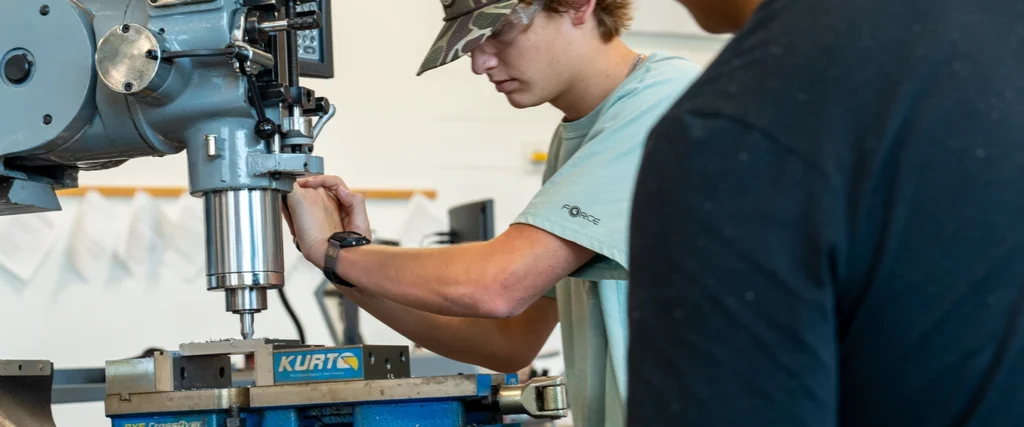 A student wearing a camouflage cap and safety glasses carefully adjusts a milling machine in a workshop setting. He is focused on machining a metal piece secured in a vise, demonstrating skills in industrial equipment operation. Another student observes closely, reinforcing a collaborative learning environment. The background features technical documents pinned to the wall and various tools, highlighting a hands-on training space designed to prepare students for careers in industrial maintenance, automation, and manufacturing systems.
