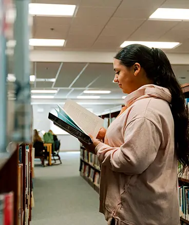 A student wearing a light pink hoodie stands in a library aisle reading a book. She is focused on the open pages, holding an additional book underneath. Bookshelves filled with books line both sides, and other people are seated and reading in the background. The space is well-lit with ceiling lights.