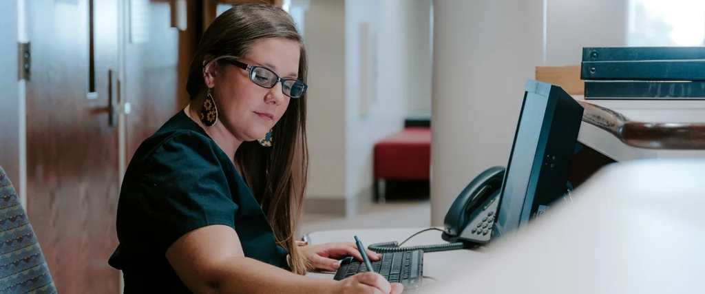 A student in the Medical Office Administration program is seated at a reception desk, attentively working on a computer while taking notes. Wearing professional attire, she demonstrates the essential skills of medical office management, including scheduling, patient communication, and record-keeping. This training prepares students for careers in healthcare administration, where organization and attention to detail are critical.