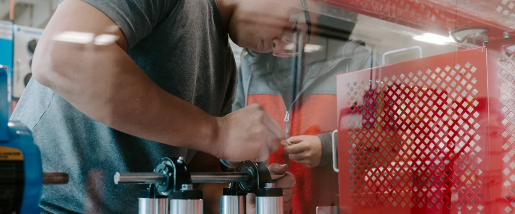 A student wearing safety glasses carefully adjusts a mechanical assembly inside a protective enclosure, demonstrating hands-on skills in mechatronics. The setup includes metal shafts, bearings, and pneumatic components, essential for understanding automation and robotics. Another student observes in the background, highlighting the collaborative nature of the learning environment. The workspace, filled with technical equipment, prepares students for careers in advanced manufacturing, robotics, and industrial automation.
