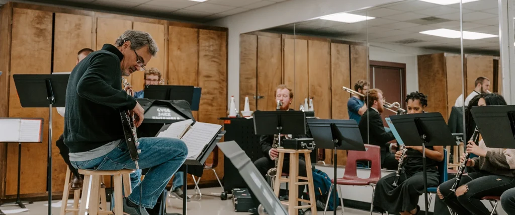 A music instructor, seated on a stool, plays an electric guitar while leading a group of students in a jazz ensemble rehearsal. The students, playing instruments like the clarinet, trumpet, and trombone, are focused on their sheet music as they perform together. The classroom features music stands, sheet music, and a mirrored wall reflecting the musicians, creating an immersive practice environment. This scene showcases the collaborative and expressive nature of a college music program.