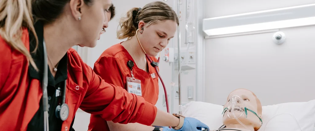 Two nursing students wearing red scrubs are engaged in a hands-on training exercise using a high-fidelity medical mannequin in a simulated hospital room. One student listens to the mannequin's vitals with a stethoscope while the other administers care, both demonstrating teamwork and clinical skills. The training environment, complete with medical equipment and monitoring systems, prepares students for real-world patient care in healthcare settings.