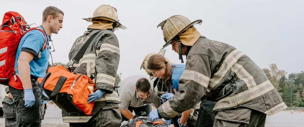 A group of emergency responders, including firefighters in full protective gear and helmets, and paramedics wearing blue gloves, are actively engaged in a rescue operation. They are gathered around a patient on the ground, preparing medical equipment and assessing the situation. One paramedic with a red backpack and another with an orange medical bag are assisting, while a firefighter leans in to help. The background shows an outdoor setting with trees, an overcast sky, and traffic cones, suggesting a training exercise or real-life emergency response.