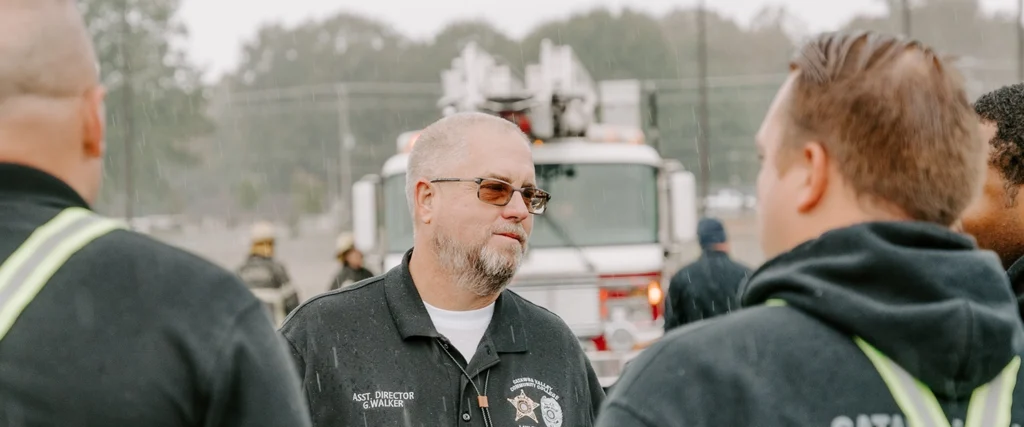 A group of emergency response professionals gather for a discussion during a rainy day. The central figure, wearing a black polo shirt with an embroidered badge and the title "Asst. Director G. Walker," listens attentively while speaking with others in reflective safety gear. Behind them, a fire truck with an extended ladder is parked, and firefighters in full gear can be seen preparing for action. Raindrops are visible in the air, adding to the atmosphere of an active emergency or training scenario.