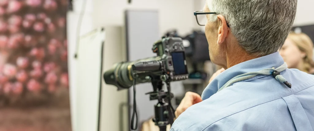 A photography instructor, wearing glasses and a light blue shirt, observes a professional DSLR camera mounted on a tripod. The camera is equipped with a telephoto lens, aimed at a subject in a well-lit studio setting. In the background, students and photography equipment, including lighting setups and backdrops, create a dynamic learning environment. The scene highlights the hands-on approach of a Photographic Technology program, where students develop expertise in camera operation, lighting techniques, and digital imaging.