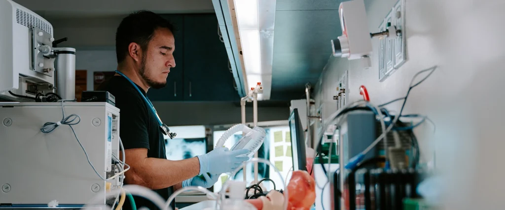 A respiratory therapy student wearing gloves and a stethoscope carefully examines a ventilator mask inside a clinical training lab. The environment is equipped with advanced medical devices, monitors, and simulation tools, providing students with real-world experience in respiratory care. This hands-on learning prepares students to assist patients with breathing disorders and emergency interventions.