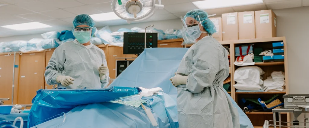 Two surgical technology students, dressed in full sterile surgical gowns, gloves, masks, and protective eyewear, stand beside a simulated patient covered in a blue sterile drape. They are in a well-equipped training lab, surrounded by medical instruments and monitoring equipment, practicing essential skills such as maintaining a sterile field, assisting in procedures, and handling surgical tools. This hands-on learning environment prepares them for roles in operating rooms, ensuring they are ready to support surgeons and patient care teams.