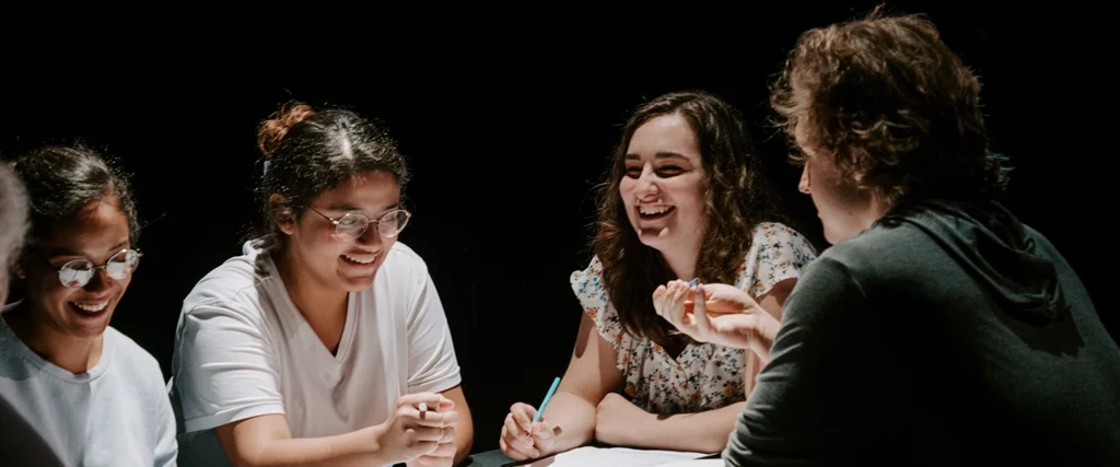 A group of theater students engage in a lively discussion during a rehearsal, illuminated by dramatic stage lighting. Their expressions convey excitement and creativity as they collaborate on a performance. The setting fosters teamwork, improvisation, and storytelling, key elements of a thriving theater program.