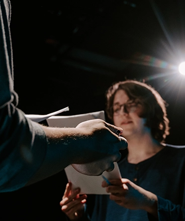 A theater student stands under dramatic stage lighting, script in hand, as they engage in a performance exercise. Another student, partially visible in the foreground, holds a script, suggesting a rehearsal or scene study. The focused expression and moody lighting capture the essence of theatrical storytelling and character development in a dynamic learning environment.