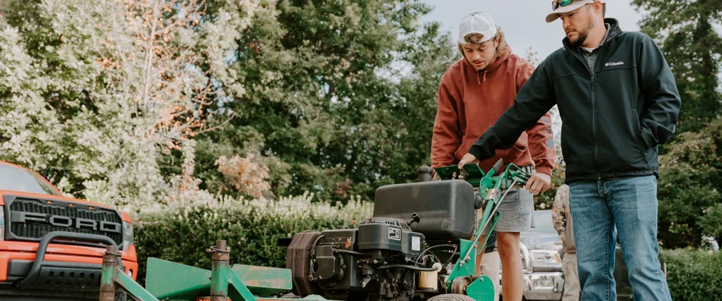 An instructor in a black jacket and sunglasses guides a student in a maroon hoodie as they examine a commercial walk-behind mower. The instructor points to the controls while the student listens attentively, preparing to operate the machine. The outdoor setting, surrounded by trees and greenery, reflects the practical, real-world training offered in a Turfgrass Management program. This hands-on experience equips students with the skills needed for careers in golf course maintenance, sports field management, and professional landscaping.