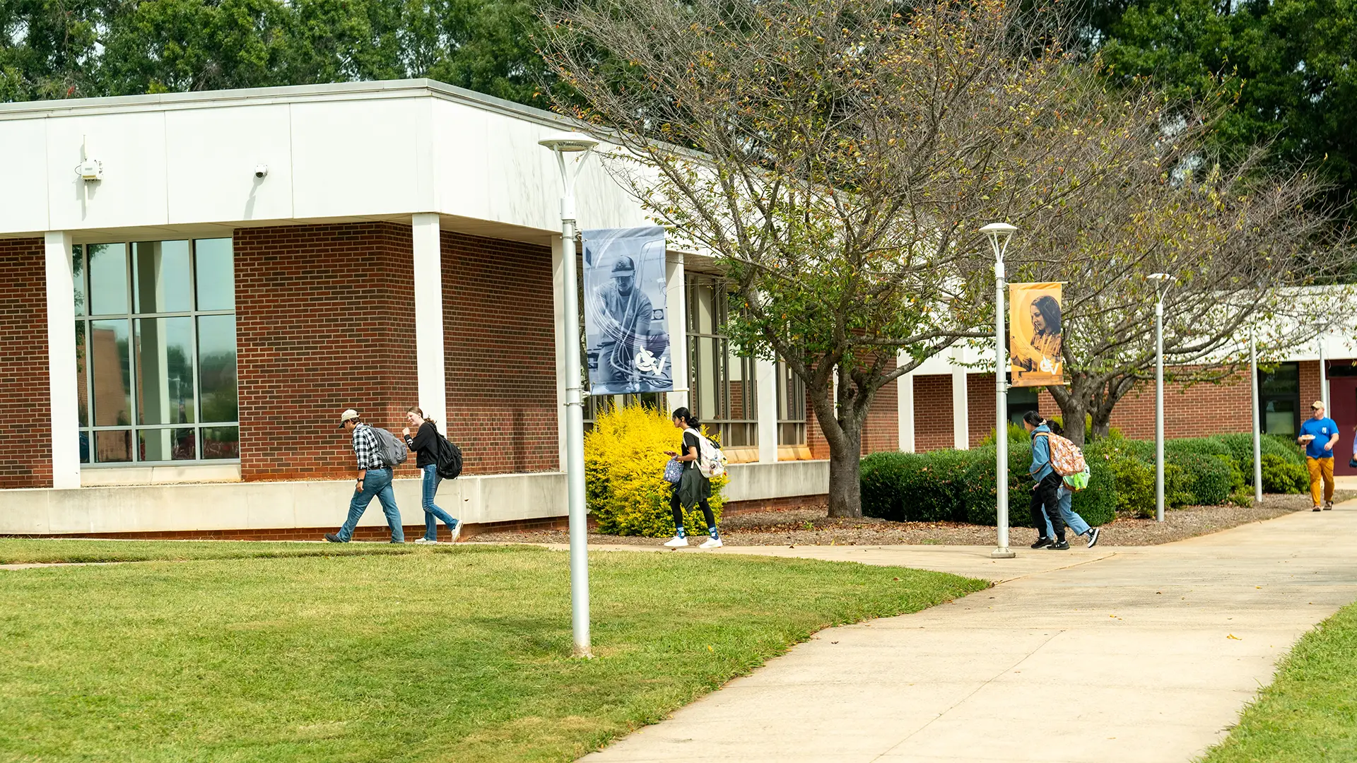 Students walk along a sidewalk past a brick and white-trimmed academic building on a college campus. Banners featuring images of students hang from lamp posts lining the walkway. Trees with sparse leaves and neatly trimmed bushes decorate the area, indicating a fall season. The atmosphere is lively with students carrying backpacks and heading toward or away from the building.