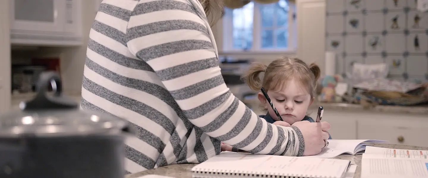 A young girl sits at a kitchen counter with a focused expression, holding a pen and working in a notebook. Beside her, an adult wearing a striped sweater assists, guiding her hand with the writing. Open books and notebooks are spread out on the counter, and a pot is visible in the foreground, indicating a multitasking parent balancing study or work and parenting.