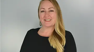 A woman with long blonde hair and light skin is smiling at the camera. She is wearing a black top and hoop earrings, standing against a simple, light gray background.