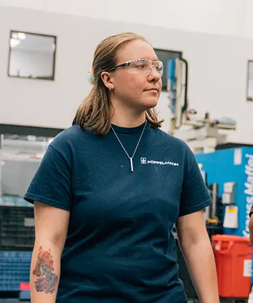 A student wearing safety glasses and a navy blue "Doppelmayr" t-shirt stands confidently in an industrial or technical training facility. She has a tattoo on her left forearm and wears a necklace. Behind her are blue machinery, a red bin, and the clean, organized interior of the workshop or lab space.