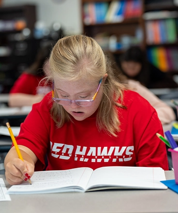 A student wearing a red “RED HAWKS” t-shirt focuses intently while writing in a notebook during class. She has blonde hair, glasses with colorful frames, and is seated at a desk surrounded by school supplies. Bookshelves and other students are visible in the background, indicating a classroom setting.