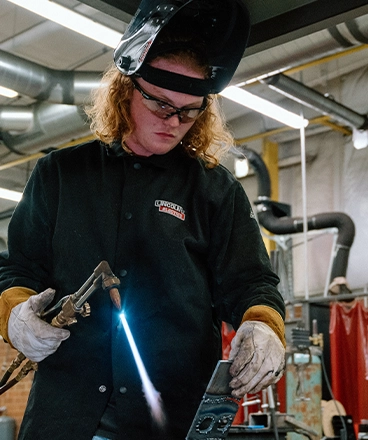 A welding student wearing protective gear, including gloves, a dark welding jacket, and a raised face shield, uses a lit torch to work on a metal piece in a workshop. The setting includes industrial equipment, ventilation pipes, and red safety curtains, indicating a hands-on training environment.