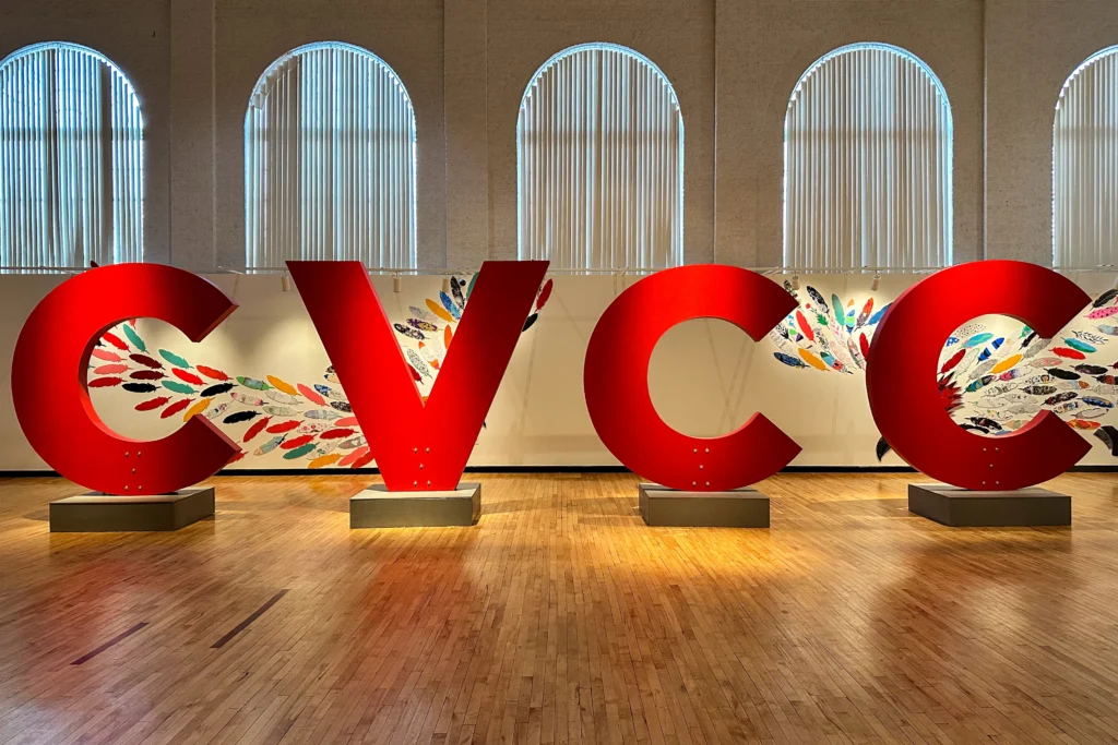 Large red letters spelling "CVCC" are displayed in a spacious gallery with arched windows and wooden floors. Behind the letters, colorful paper feathers form wings on the wall, creating a vibrant and artistic backdrop.