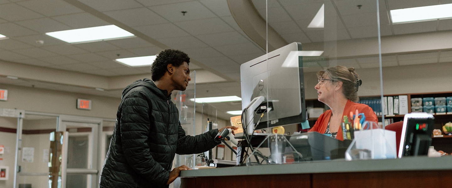 A student in a black jacket speaks to a staff member at the library front desk. The staff member, seated behind a plexiglass barrier, is wearing glasses and a red cardigan. A desktop computer and office supplies are visible on the desk in a well-lit library setting.